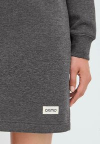 Gray sweatshirt dress with a soft texture, featuring a visible logo tag at the hem. The design includes long sleeves and a relaxed fit.