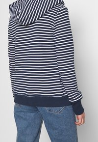 Felpa a righe blu navy e bianche con cappuccio regolabile e polsini a costine, abbinata a jeans azzurri chiari, che mostrano una vestibilità rilassata e uno stile informale.