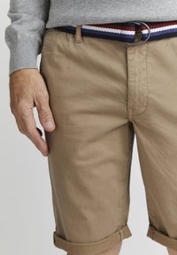 Beige bomullsshorts med rullekantar, med eit navyt blått, raudt og kvitt vevd belte. Hand som kviler på sida, som viser stoffteksturen.