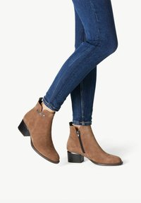 Stivaletti in suede marrone con tacco basso a blocco, zip laterale e accento metallico sulla parte superiore. Indossati con jeans skinny di denim scuro.