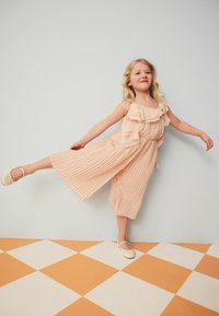 Jumpsuit en pêche plissé avec des bretelles volantées et des chaussures douces assorties. La tenue présente des rayures verticales et une taille cintrée pour un ajustement parfait.