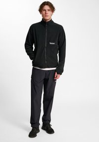 Svart fleecejacka med ståkrage och dragkedja framtill, i kombination med mörka byxor och svarta sneakers. Märkbar logotyp på jackfickan.