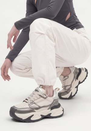 Person iført hvide joggebukser og grå langærmet top sidder i knæ, og viser chunky hvide og grå sneakers med tykke sorte såler.