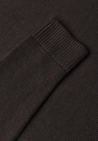 Manche de pull en maille brun foncé avec bord-côte sur un fond de tissu texturé assorti.