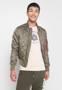 Olivgrüne gesteppte Bomberjacke mit gerippten Bündchen und rotem Etikettenakzent, getragen über einem hellen T-Shirt, kombiniert mit olivgrünen Jogginghosen.