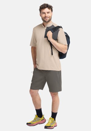 Beige kurzärmeliges Polo, graue Shorts und gelbe Sneakers mit schwarzen Akzenten. Das Model hält einen schwarzen Rucksack und steht in einer neutralen Pose.