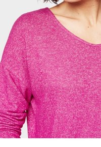 Kobieta nosząca jasnoróżowy sweter z długim rękawem i okrągłym dekoltem, ukazana od ramion do połowy torsu.