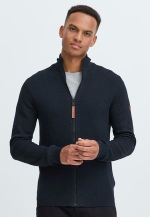Mornarški zip-up pulover iz strukturirane pletenine. Ima visok ovratnik ter rebraste manšete in spodnji del. Usnjeni dodatek blizu rokava.