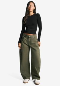 Žena stojaca v priliehavom čiernom crop top s dlhým rukávom, širokých olivovo zelených nohaviciach a bielych teniskách na bielom pozadí.