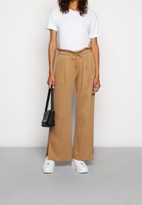 Beżowe spodnie dresowe o szerokich nogawkach, z wiązaniem w pasie, bocznymi rozcięciami i białym logo Nike. W komplecie z białą koszulką i białymi sneakersami.