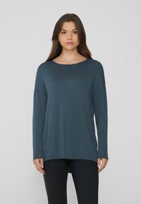 Dunkelblau-grünes Langarmoberteil mit rundem Halsausschnitt, lockerer Passform und angeschnittenen Schultern. Glatte Textur, kombiniert mit schwarzen Leggings.