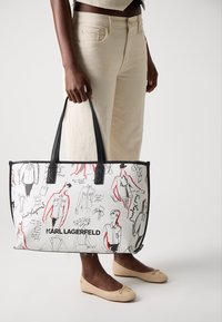 White tote bag with black and red sketches, featuring a black logo at the base. Smooth material, rectangular shape, dual shoulder straps.