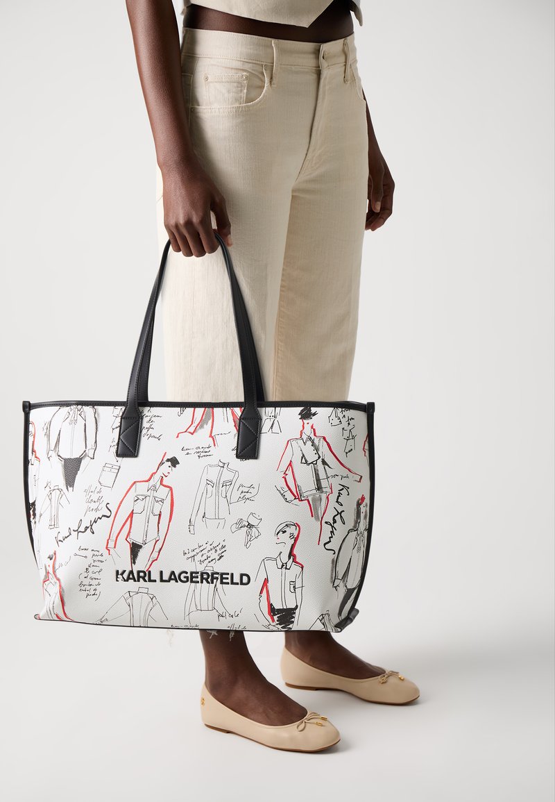 White tote bag with black and red sketches, featuring a black logo at the base. Smooth material, rectangular shape, dual shoulder straps.