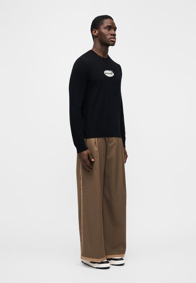Black sweater with a round neck and "Voltaire" logo on the chest, paired with loose beige pants and black-and-white shoes. Simple design.