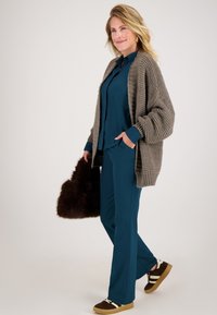 Teal blouse en bijpassende broek gecombineerd met een chunky, textuur grijze cardigan. Houdt een donkerbruine leren tas vast, met bruine sneakers met witte strepen.