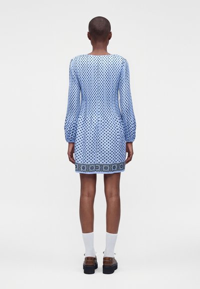 Personne aux cheveux courts, vue de dos, portant une robe bleue à manches longues avec un motif à pois et un ourlet décoratif, des chaussettes blanches et des chaussures marron.