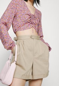 Cropped top floreale in rosa, giallo e viola con maniche lunghe e dettaglio a fiocco. Pantaloni corti beige a vita alta con tasche laterali e cintura. Borsa rosa.