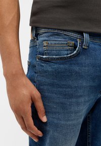 Blå denimjeans har en klassisk femfickdesign med synliga sömmar och en något blekt textur. En hand vilar på sidan.