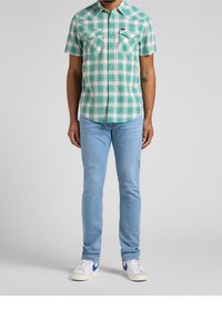 Karierte Kurzarmbluse in Türkis und Weiß, mit zwei Brusttaschen und einer geknöpften Leiste. Kombiniert mit hellblauen Jeans und weißen Sneakern.