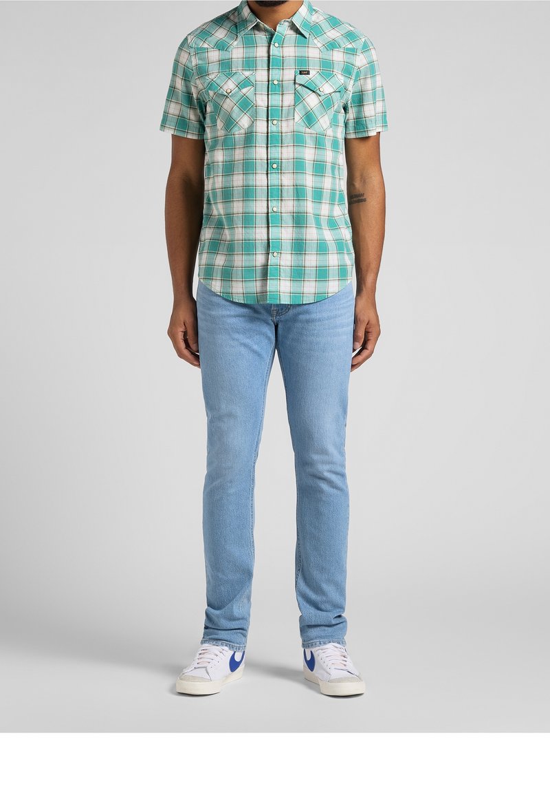 Karierte Kurzarmbluse in Türkis und Weiß, mit zwei Brusttaschen und einer geknöpften Leiste. Kombiniert mit hellblauen Jeans und weißen Sneakern.