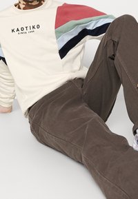 Felpa crema con pannelli multicolori in rosso, verde, blu e blu scuro; presenta il logo "KAOTIKO". Pantaloni in corduroy marrone con una texture sottile.