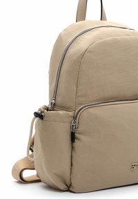 Beige stoffen rugzak met afgeronde hoeken, twee ritssluitingvakken, een voorvak en verstelbare riemen met logo-accent.