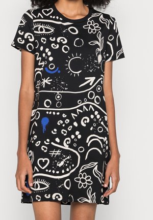 Černé krátké šaty s bílými abstraktními tvary, květinami, očima a šipkami, doplněné malými modrými akcenty, nošené stojící osobou.