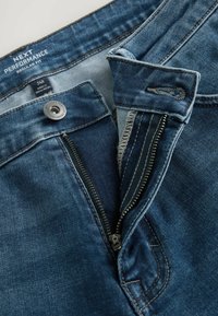 Jeans de denim azul con un ajuste regular, que cuentan con una cremallera, botón metálico y costuras en contraste. Etiqueta visible en la parte interior de la cinturilla.