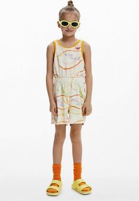 Hvid heldragt med orange og grønne blomsterprint, elastisk talje, kombineret med gule solbriller, orange sokker og gule sandaler.