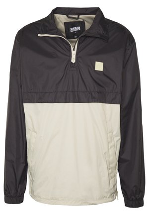 Mustan ja beeži üleminekujakk, valmistatud kergest kangast, millel on veerand-pikkuses tõmblukk, elastsed mansetid ja väike logo pilt rinnal.