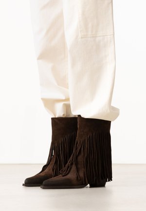 Bruine suède enkellaarsjes met franjes, gedragen met losse witte broek tegen een effen achtergrond.