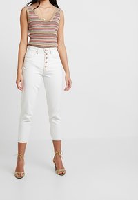 Randig stickad tanktopp med V-ringning, kombinerad med vita högmidjade jeans med knappgylf och ankellängd. Guldskor framhäver outfiten.