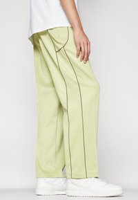 Light green, loose-fitting pants made of soft fabric, featuring contrasting brown stitching and a straight cut. Worn with white sneakers.