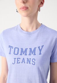 Šviesiai levandų spalvos medvilnės marškinėliai, su išblukusiu mėlynu grafiniu užrašu "TOMMY JEANS" priekyje ir klasikiniu apvaliu kaklu.