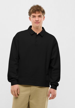 Young man with short curly hair wearing a black long-sleeve polo shirt and beige pants, standing against a plain white background.