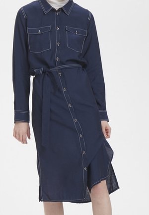 Kvinde iført en navyblå midi-kjole med knapper, hvide syninger, brystlommer og bælte i taljen, stående foran en ensfarvet baggrund.