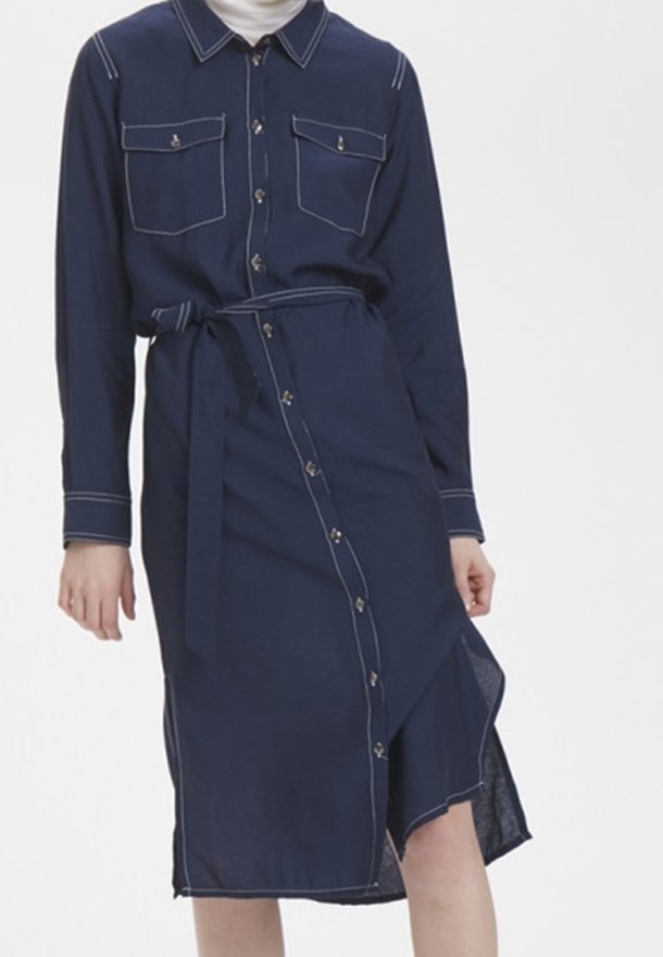 Femme portant une robe midi boutonnée bleu marine avec des surpiqûres blanches, des poches poitrine et une ceinture à la taille, posant devant un fond uni.