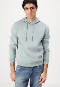 Sweatshirt verde claro com bolso frontal e capuz ajustável, feito em tecido suave. Combinado com jeans azuis.