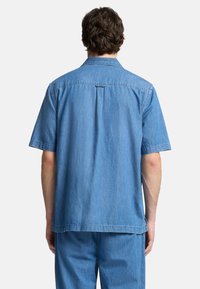 Camicia di jeans a maniche corte di colore blu chiaro con taglio dritto, piega sul retro e colletto semplice. Il tessuto presenta una finitura morbida e testurizzata.