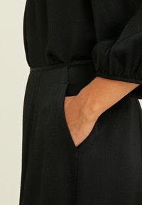 Schwarzes Stoffkleid mit strukturiertem Oberflächen, einer definierten Taille mit Seitentasche und sanft gepufften Ärmeln.