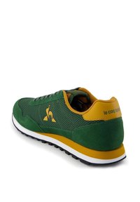 Scarpa sportiva verde in materiale mesh e scamosciato, con dettagli gialli, suola bianca e logo sul tallone. Presenta un design con lacci frontali.