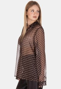 Camicia trasparente marrone con pois bianchi, chiusura a bottoni frontale, maniche lunghe e una tasca sul petto.