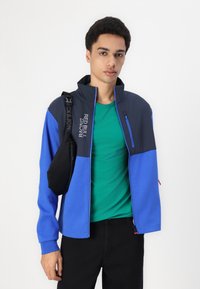 Modra in črna zip-up jakna z visokim ovratnikom, pod katero je vidna zelena majica. Črna torbica je nosena čez ramo.
