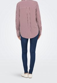 Personne portant une chemise rayée verticale rouge et blanche à manches longues, un jean skinny bleu foncé et des chaussures blanches à enfiler, debout dos face à l'avant.