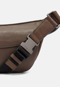 Brązowa skórzana torebka typu crossbody z teksturowanym wykończeniem, regulowanym paskiem z metalowym zapięciem oraz zaokrąglonym kształtem.