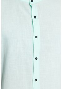 Camicia verde menta chiara realizzata in un tessuto texturizzato, con chiusura a bottoni neri sul davanti. Il colletto è rotondo e presenta cuciture a contrasto.