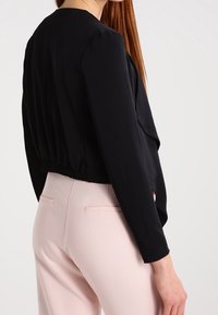 Veste noire courte avec des manches longues, présentant un devant drapé et un ourlet élastique. Associée à un pantalon rose clair, mettant en avant une texture lisse.