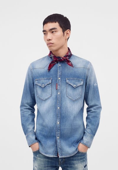 Young man wearing a faded blue denim shirt and jeans with hands in pockets and a red plaid neckerchief tied around his neck.