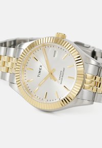 Montre Timex avec un bracelet en acier inoxydable, lunette plaquée or, cadran argenté, index horaires et dateur ; étanche jusqu'à 50 mètres.