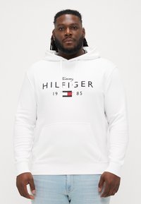 Biela mikina s predným vreckom a sťahovacou kapucňou. Obsahuje čierny nápis "Tommy HILFIGER" a logo v červenej, modrej a bielej farbe. Mäkká textúra látky.